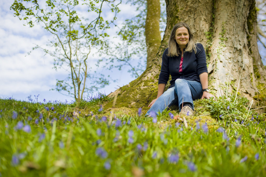 Audrey sitting against a large tree with grass and bluebells in the foreground.